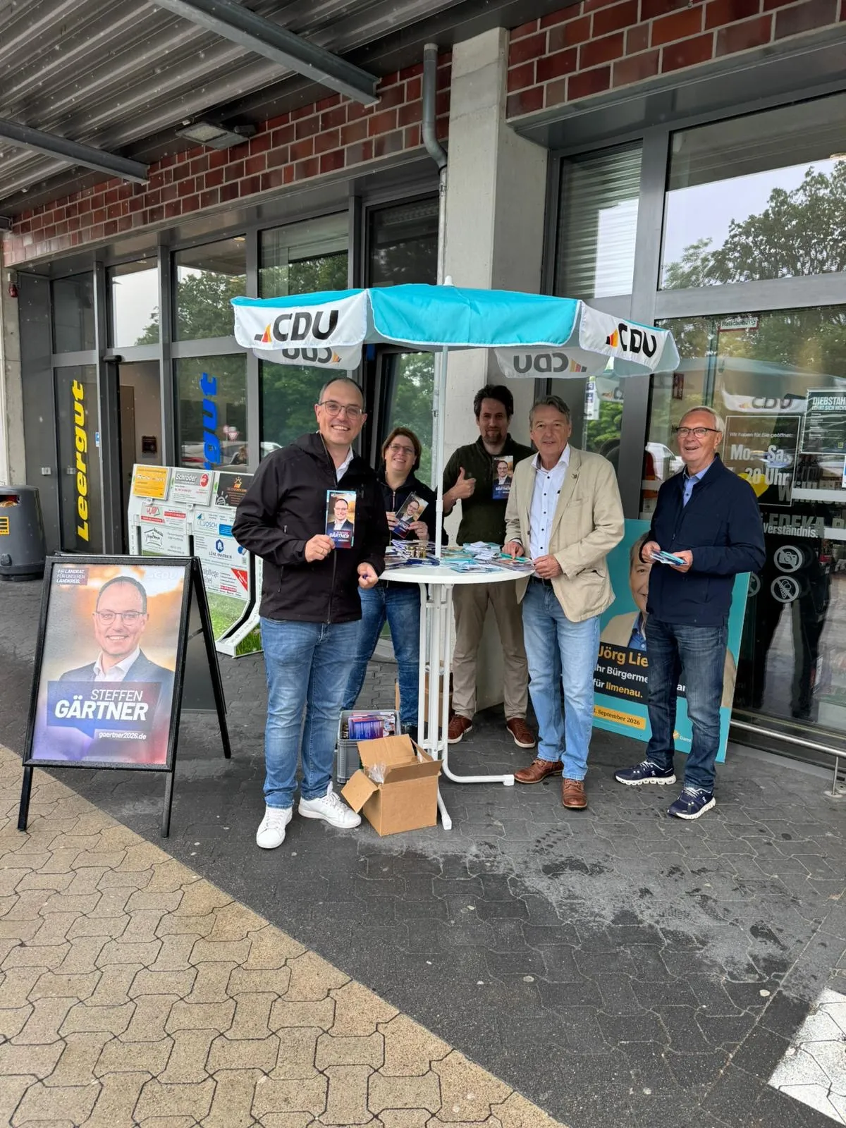 Das Team des CDU Samtgemeindeverbands Ilmenau am Infostand unter einem CDU-Sonnenschirm vor dem EDEKA in Melbeck, links ein Plakat von Landratskandidat Steffen Gärtner.